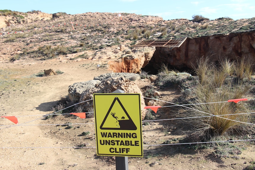 A yellow sign reading 'warning unstable cliff' with a barricade and coastal hole behind