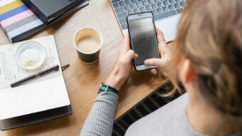 A woman uses her phone in front of a laptop while drinking a coffee.