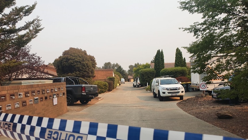 Police tape appears out the front of a driveway near houses. A police car is parked there.