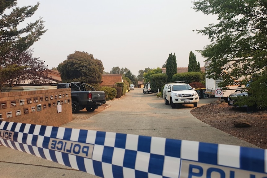 Police tape appears out the front of a driveway near houses. A police car is parked there.