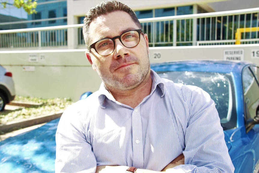 A man in a business shirt leans against a car in the sun.