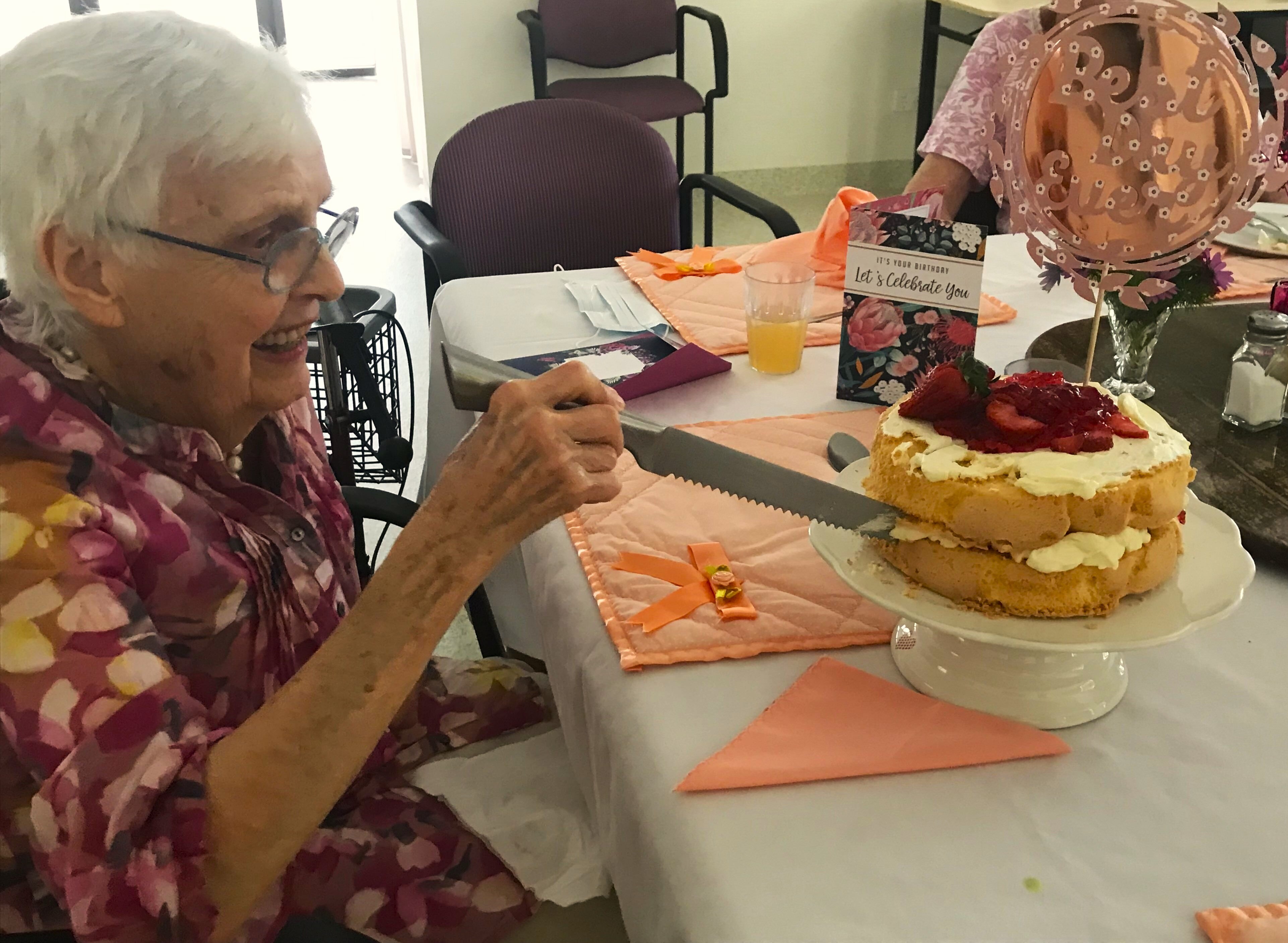An elderly woman cuts a piece of a strawberry and cream birthday cake