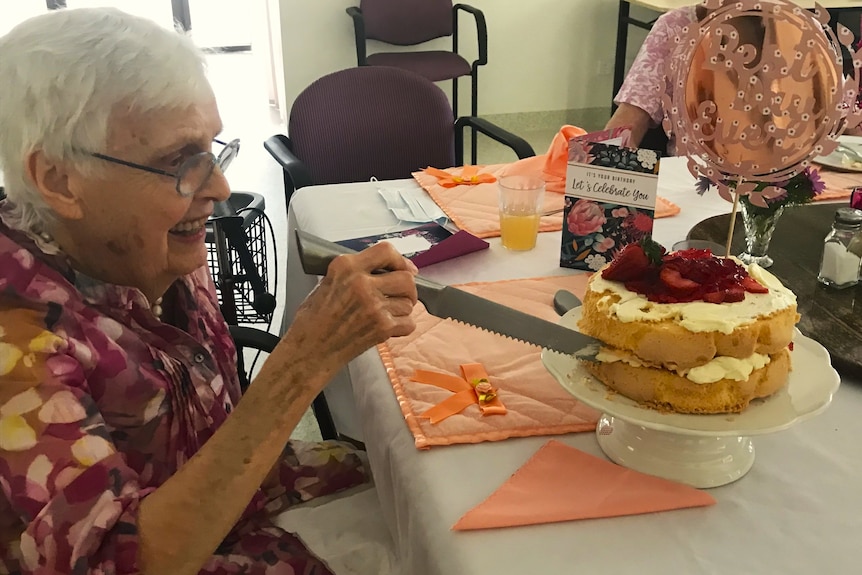 An elderly woman cuts a piece of a strawberry and cream birthday cake