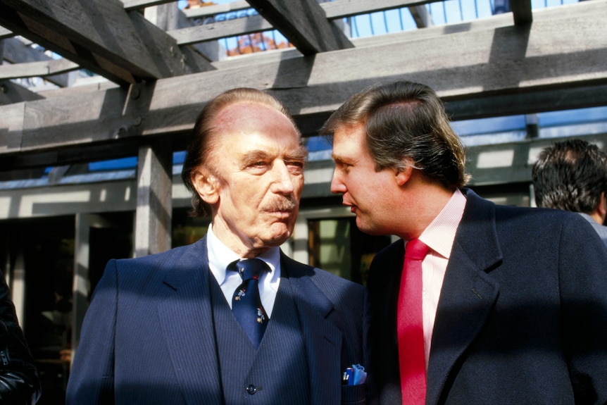 A young Donald Trump, wearing a black suit and red tie, leans in to speak with his father Fred, a mustachioed man in blue suit