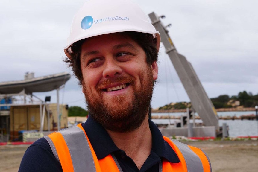 A young man with a scrubby beard, wearing a hard hat smiling at something off-camera.
