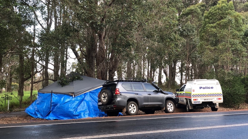 A tent with a tarpaulin pulled around it protects the scene. A black 4wd and police car are parked nearby.