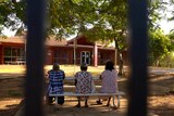 Three aboriginal women are seen through a fence sitting at a table facing a remote hospital.