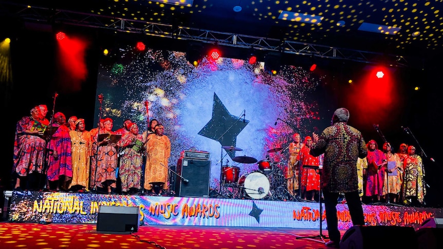 Central Australian Aboriginal Women's Choir stand on a colourful, low stage in two groups in patterned dresses, with conductor