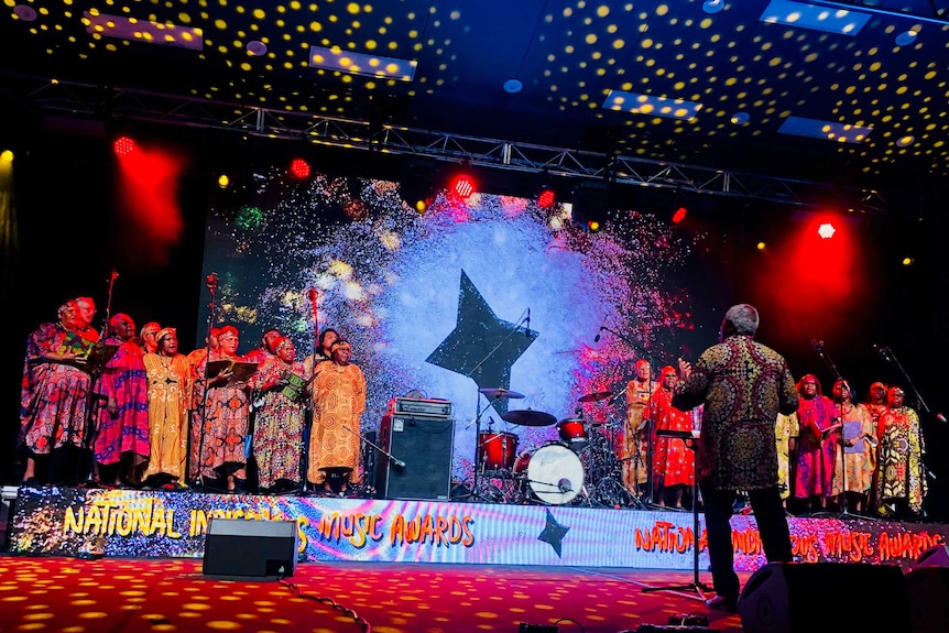 Central Australian Aboriginal Women's Choir stand on a colourful, low stage in two groups in patterned dresses, with conductor