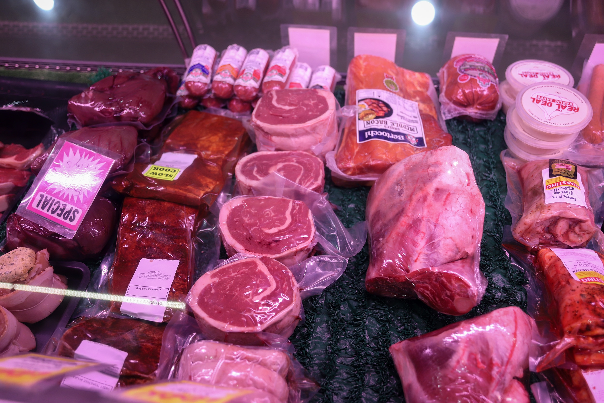 A display of a range of cuts of meat under glass and pink light in a butchers shop.