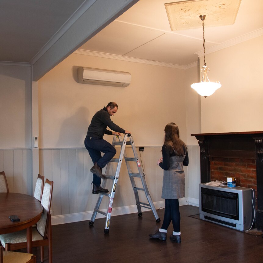 A man climbs down a ladder inside a living room, with an air conditioner on the wall, gas heater on the floor