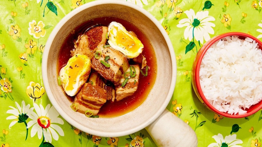 Pork and eggs in a brown braising sauce in white claypot next to dish of rice on a colourful green flower tablecloth. 