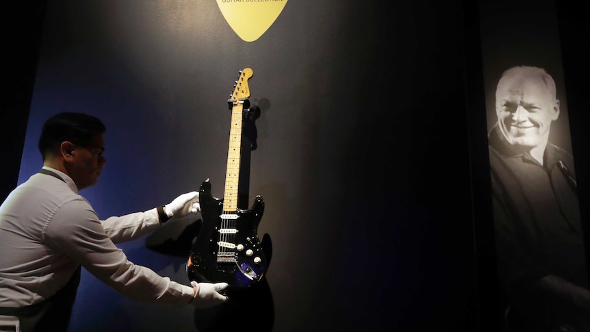 A man wearing gloves carefully places a shiny black guitar on a wall for display.