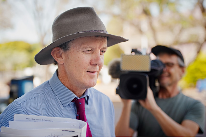 A man wearing a dress shirt, tie and wide-brimmed hat walks carrying a bundle of papers. 