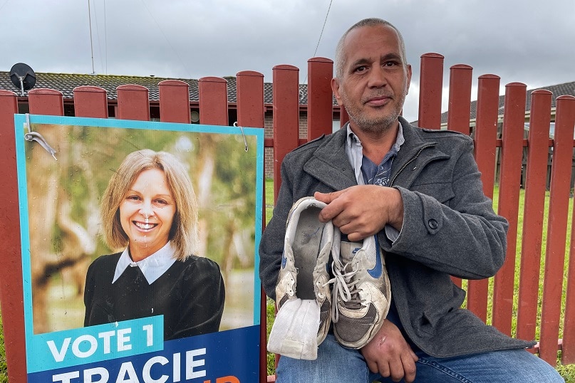 A man sits next to a political corflute holding his joggers missing a shoelace.