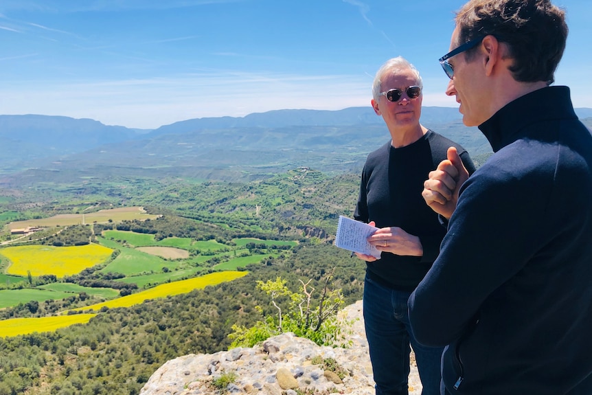 Man with white hair, sunglasses, black jumper and notebook looks at brown hair man standing over picturesque valley of vineyards