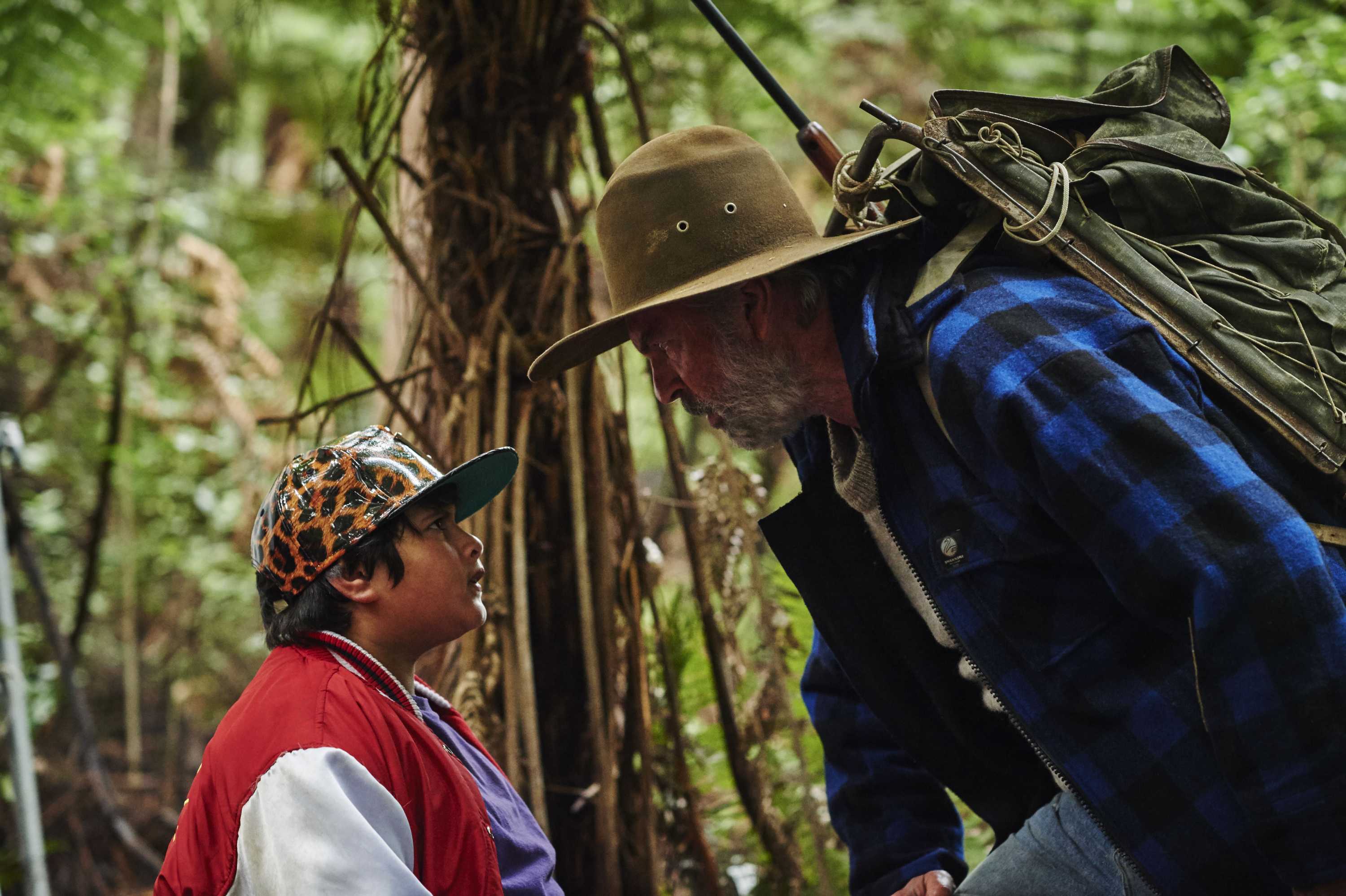 Sam Neill in an akubra hat leans down towards a young Julian Dennison, they are in the bush