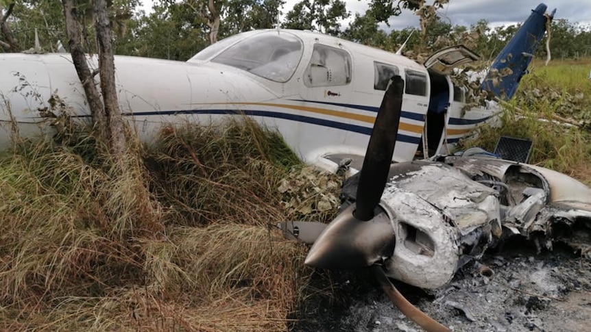 A wrecked white plane with yellow and blue stripes lies on the grass.