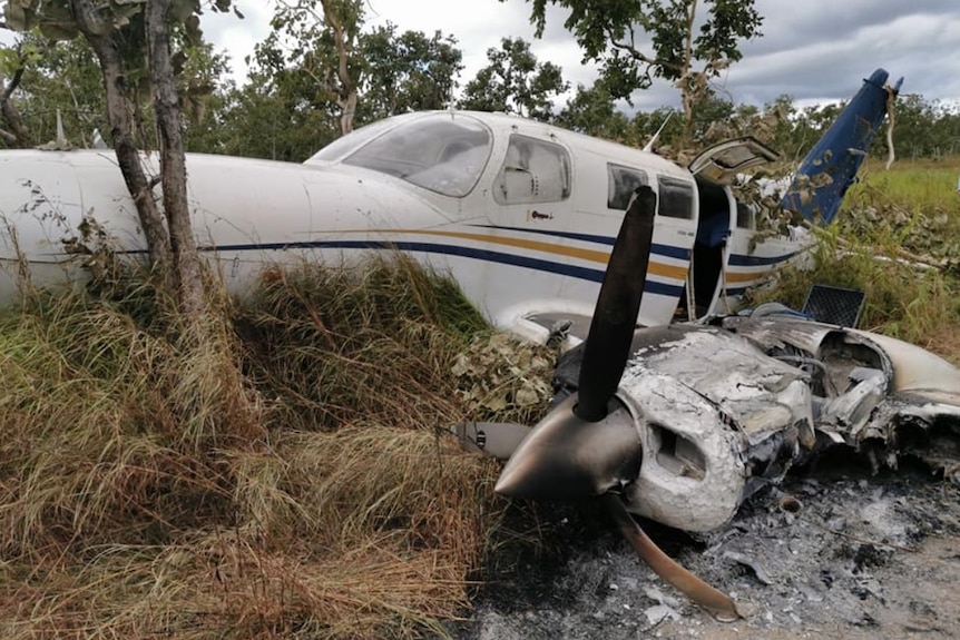 A wrecked white plane with yellow and blue stripes lies on the grass.