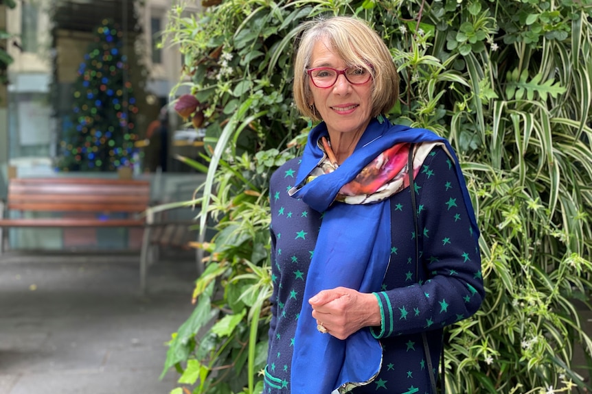 A woman wearing a blue scarf and red glasses stands in front of a wall of greenery, smiling