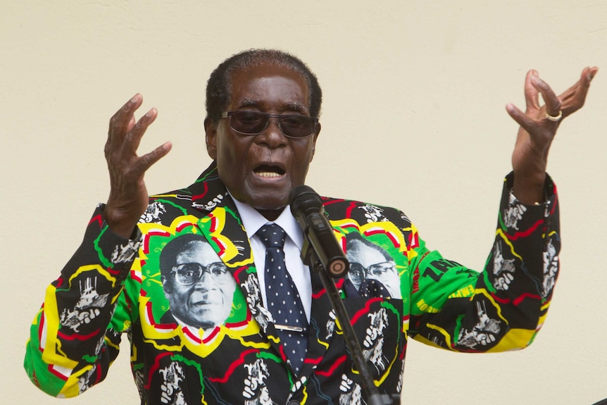 Robert Mugabe wears a colourful jacket featuring photos on his own face as he speaks at an event and gestures with his hands.