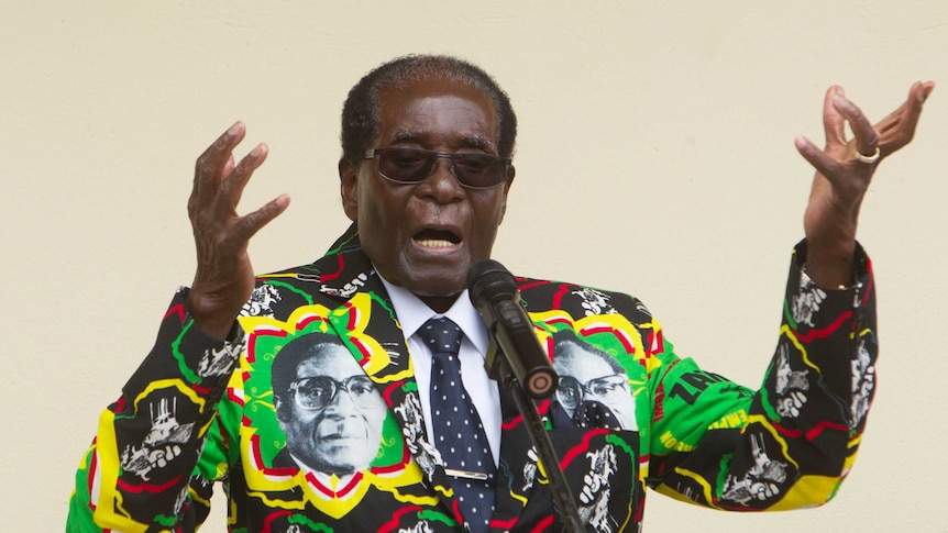 Robert Mugabe wears a colourful jacket featuring photos on his own face as he speaks at an event and gestures with his hands.