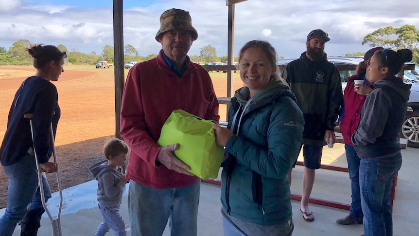 A lady in a green jacket hands over a fluro yellow bag to a man in a red jumper