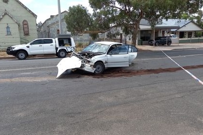 A white car with a badly damaged bumper with its doors open sitting at the end of a skid on a road