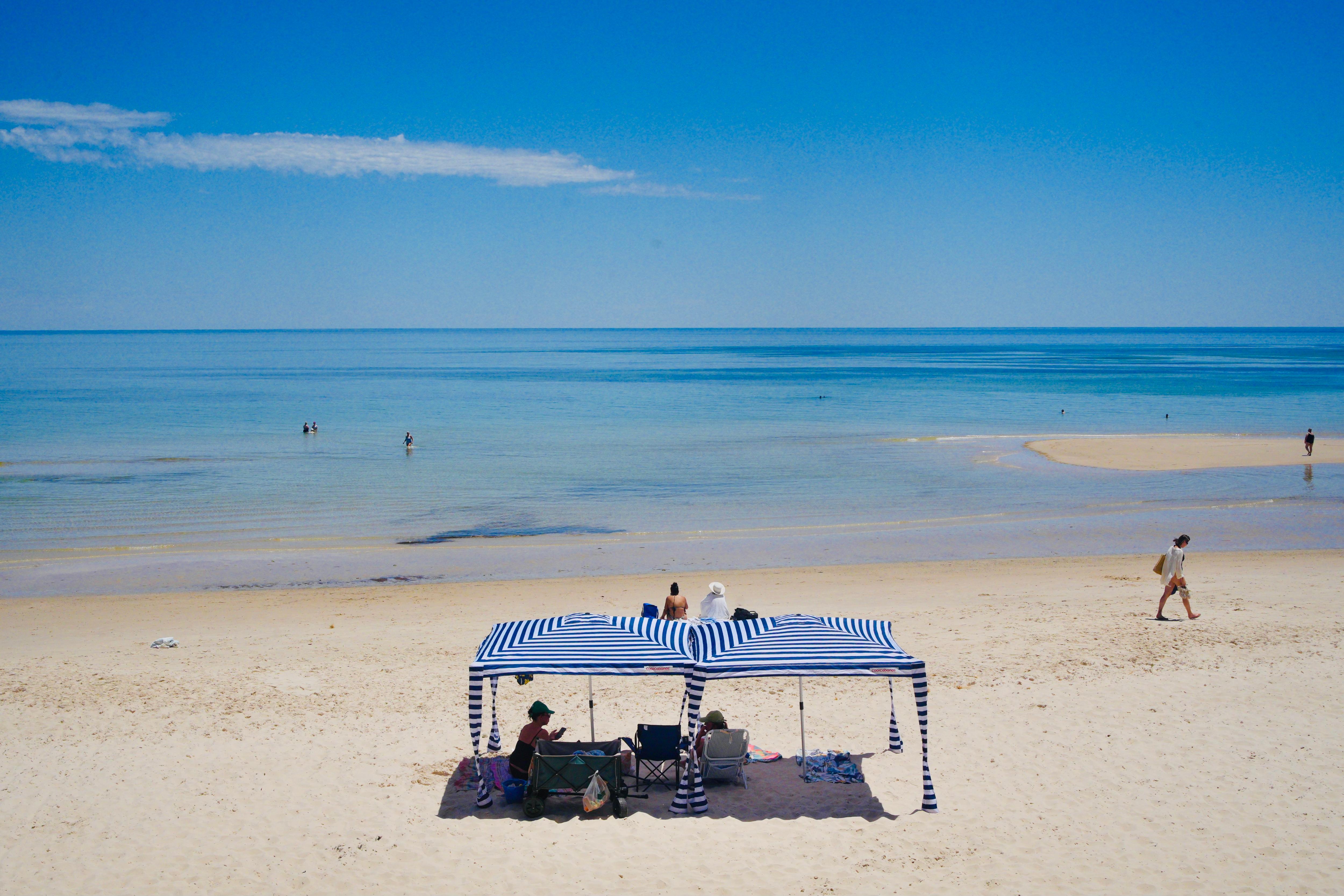 Alguns banhistas ficam à sombra de uma cabana listrada de azul e branco em um trecho de praia. 