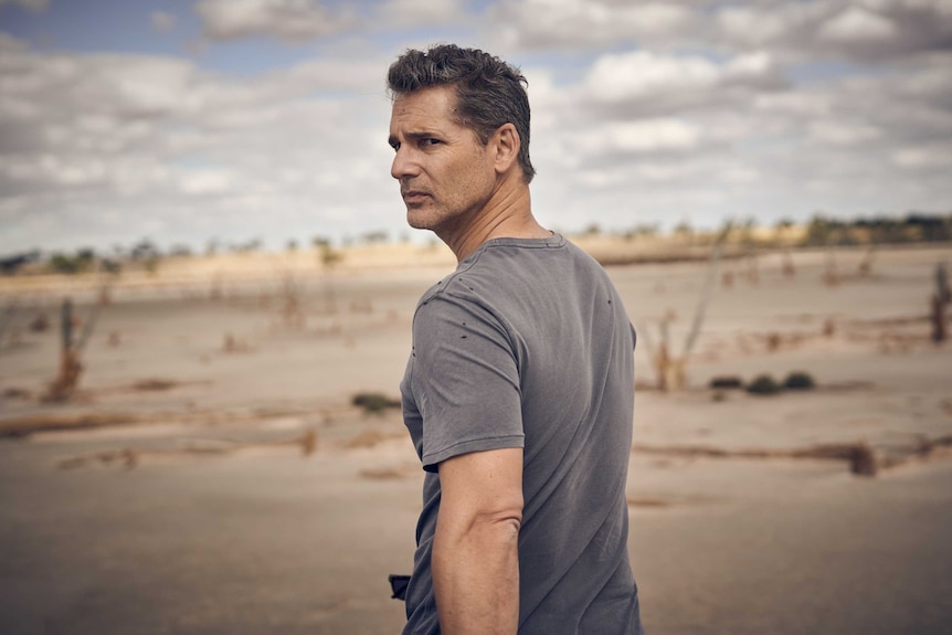 A dark-haired man in a washed out dark grey t-shirt stands in a drought stricken plain side-on