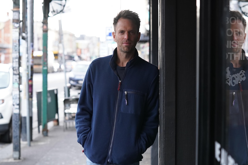 Man in a navy zip-up jumper leans with crossed arms against a rendered wall on a Melbourne street 