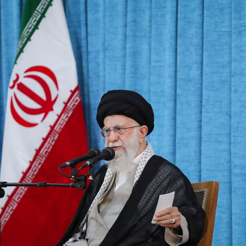 A bearded man in a turban and glasses speaks into a microphone in front of the Iranian flag.