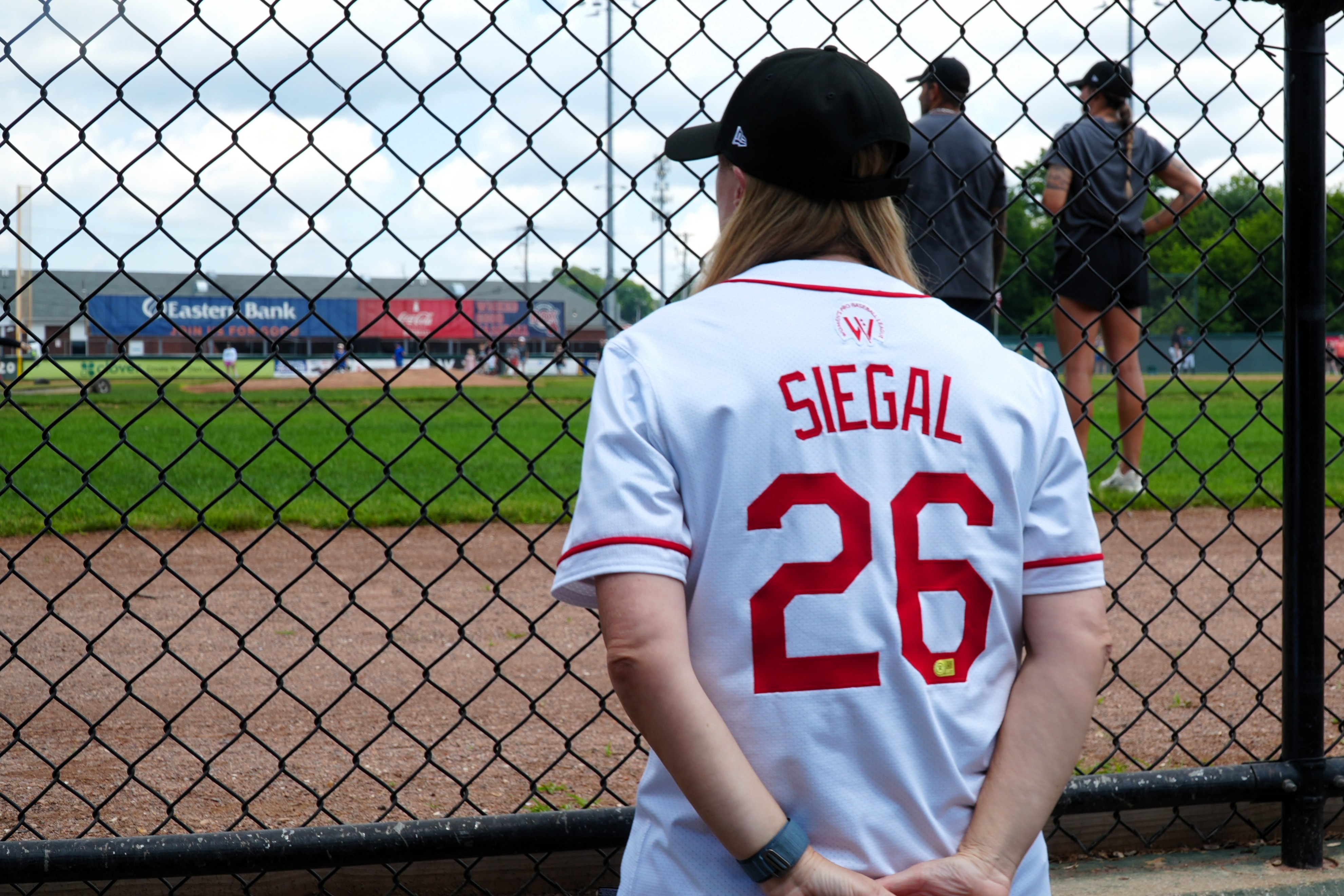 A shot from behind of a woman watching baseball training, she wears a jersey that says 'Siegal'