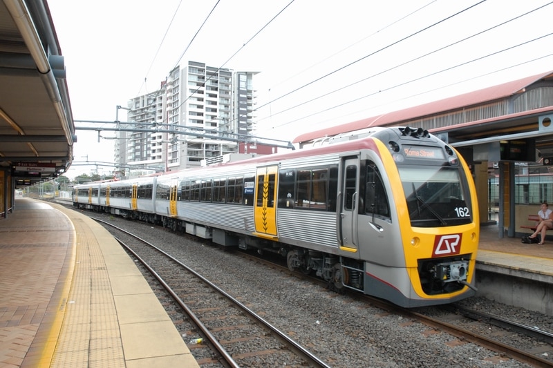 A grey and yellow electric train pulls into a an inner-city train station.