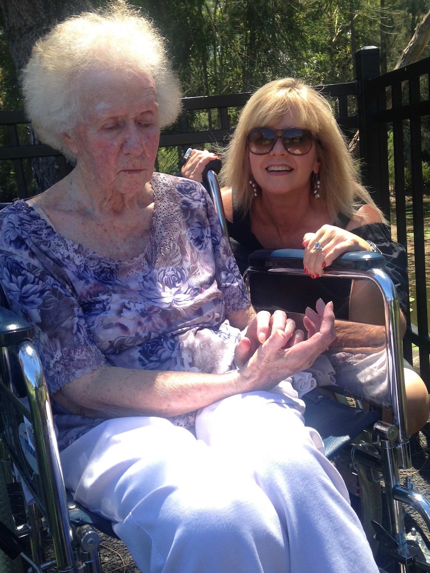 An elderly woman sits in a wheelchair, looking at her hands. A younger woman next to her smiles.