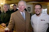 Prince Charles holds a glass of beer at the bar of a pub, standing next to a smiling man.