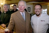 Prince Charles holds a glass of beer at the bar of a pub, standing next to a smiling man.