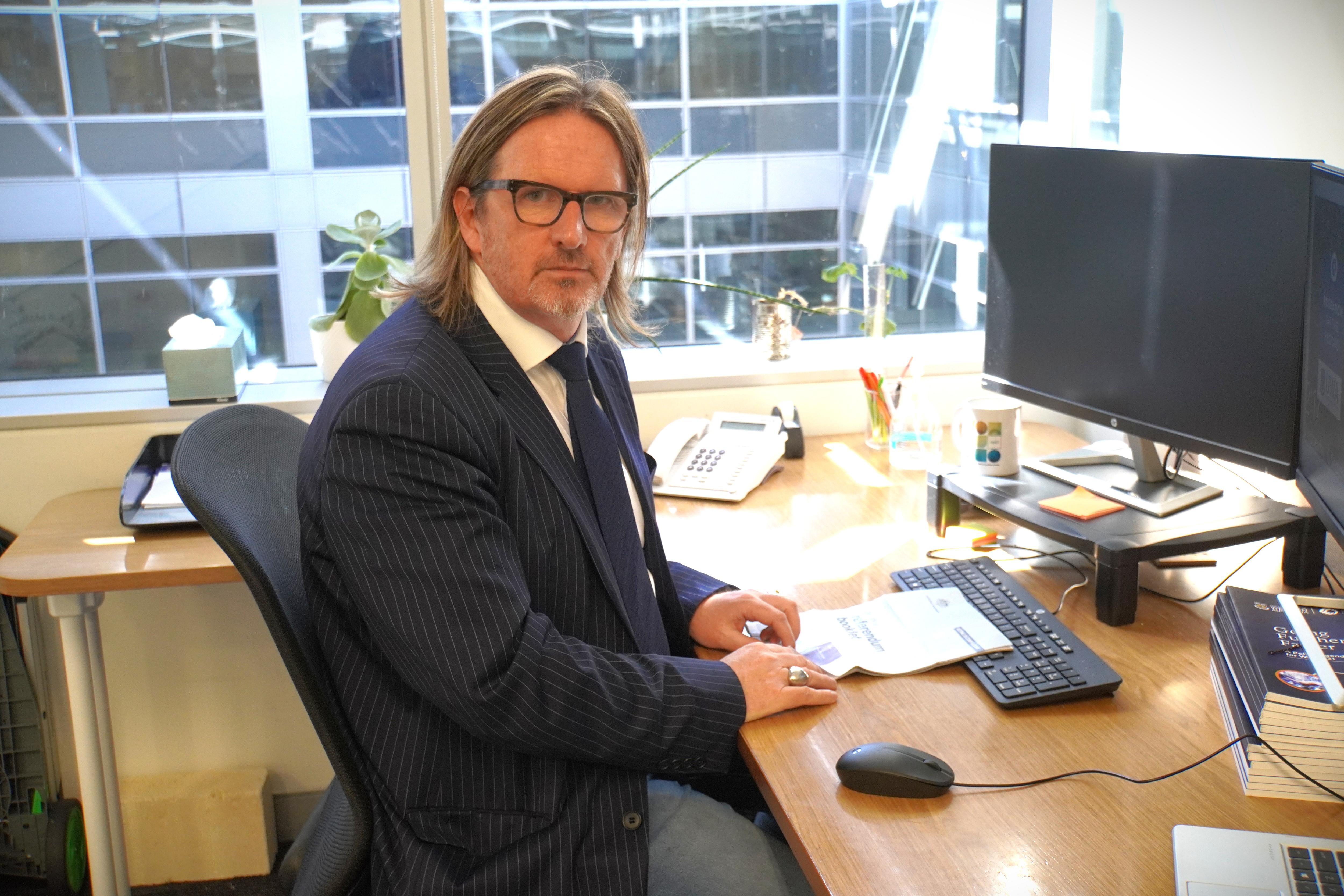 A middle-aged man sits at a desk and computer in an office with his face turned sideways towards the camera.