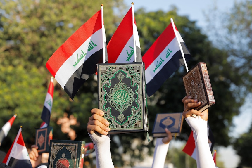 A close-up photo shows a green and gold book being held in the air in someone's hand, in front of Iraqi flags at a rally.