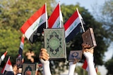 A close-up photo shows a green and gold book being held in the air in someone's hand, in front of Iraqi flags at a rally.