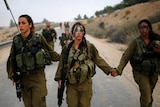 A group of young female Israeli soldiers walk down a road, looking tired. Two are holding hands