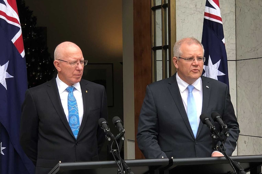 Two men in suits stand behind podiums decorated with the Australian national emblem with other people present