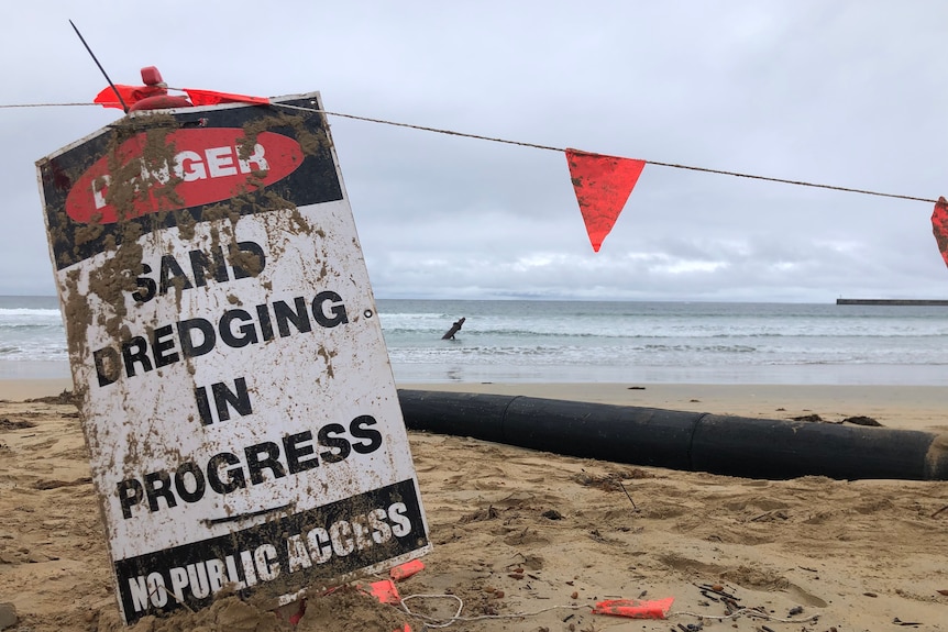 A white and black sign with red danger on it and  'sand dredging in progress - no public access' on a beach.