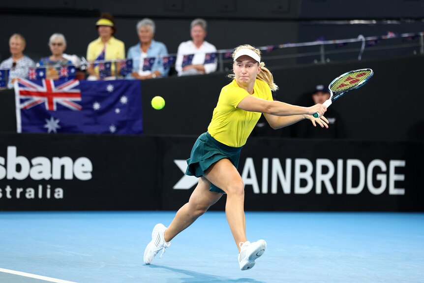 Daria Saville stretches to play a backhand while wearing a yellow top and green skirt. Fans hold Australian flags behind her.