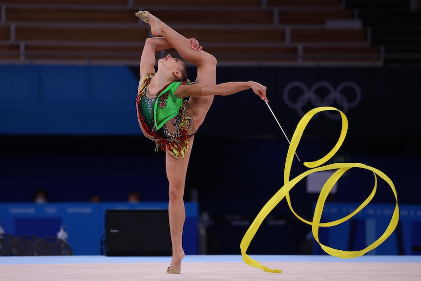 A rhythmic gymnast stands on one tiptoe while holding her other leg behind her back, waving a ribbon. 