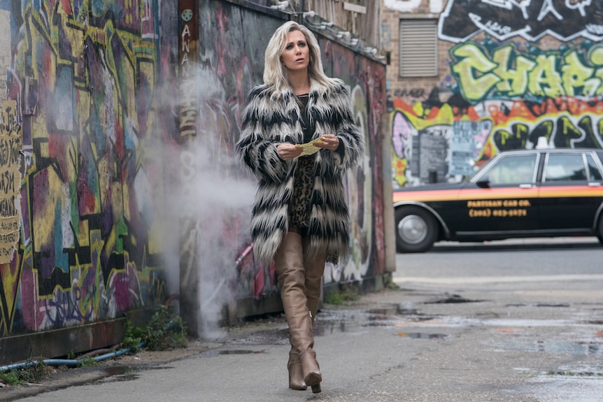 Kristen Wiig wearing ratty black and white shaggy coat and thigh-high beige boots, walking in graffiti lined laneway.