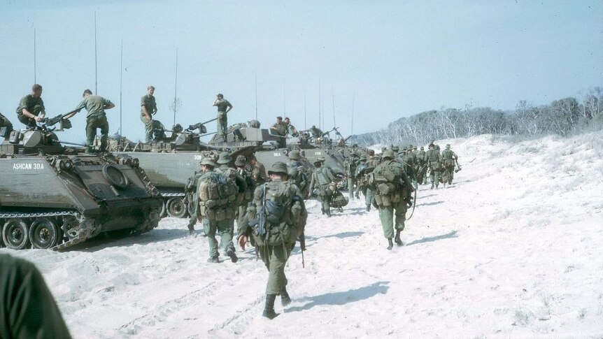 Australian troops carry their packs walking past a line of tanks lined up on a beach.