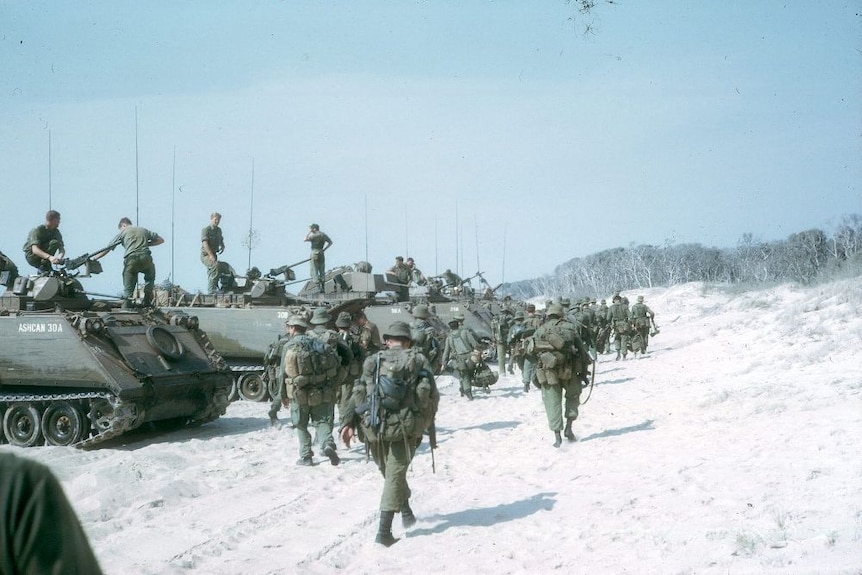 Australian troops carry their packs walking past a line of tanks lined up on a beach.