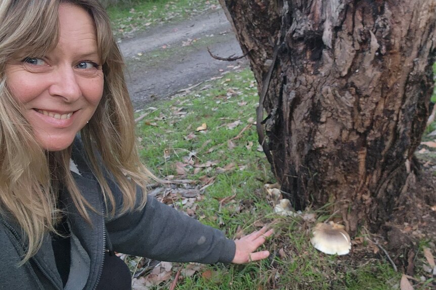 A woman with long blonde hair crouches down on the grass, pointing to a small mushroom at the stump of a tree.