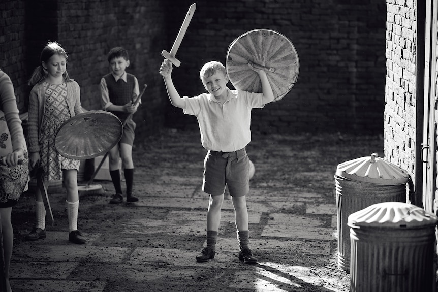 A black and white image of a boy in 60s-style clothing grinning as he holds up a toy sword and shield